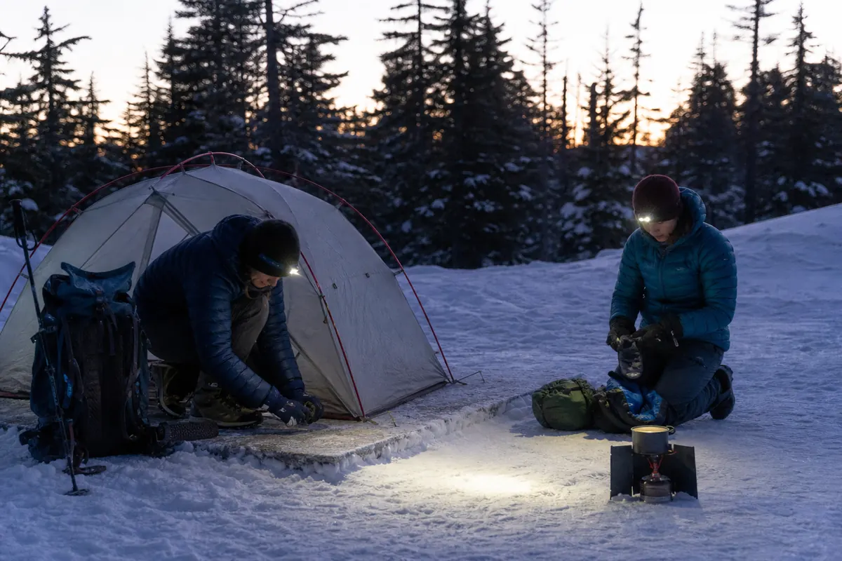 Winter backpackers following a shelter-first camp sequence with warm layers, controlled snow platform, stove, and protected dry gear - winter camp setup | KnowOutdoor