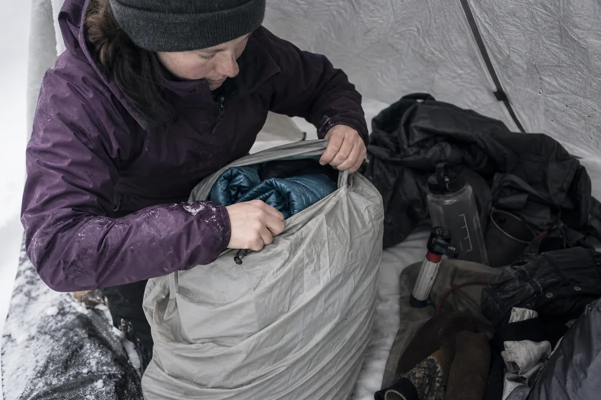 Winter backpacker sealing sleeping insulation, dry base layers, and spare mittens inside a waterproof pack liner away from snowy gear - winter dry insulation | KnowOutdoor