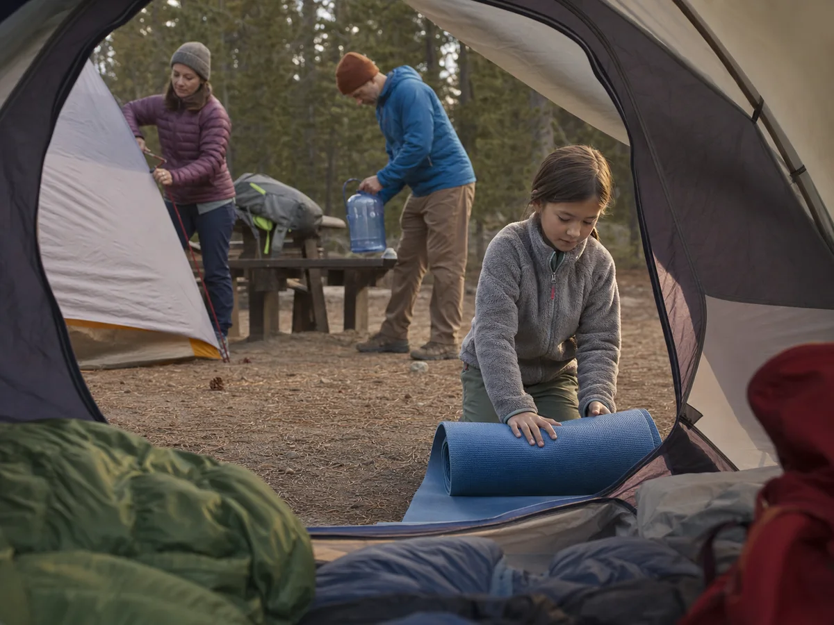 Child unrolling a sleeping pad inside a small family backpacking tent while adults manage shelter, water, and food storage at a generic campsite - family camp routine | KnowOutdoor