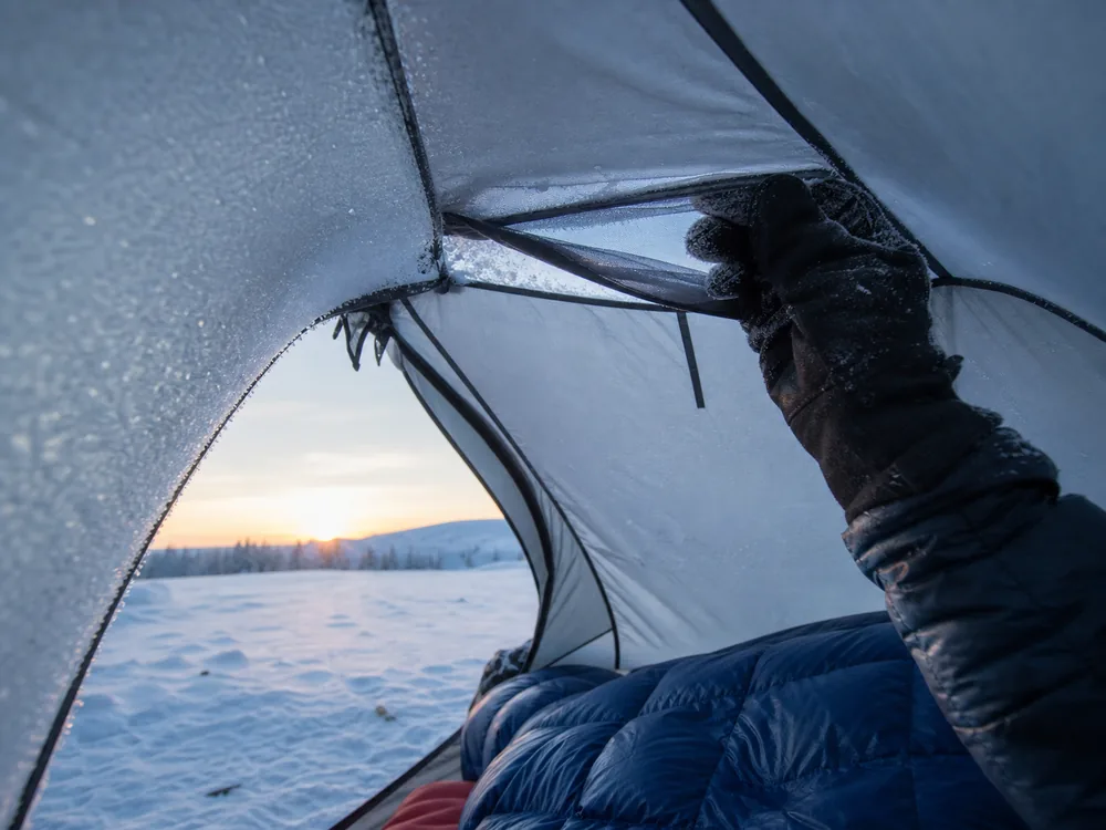 Winter camper clearing frost from an open tent vent while keeping a sleeping bag away from the wall
