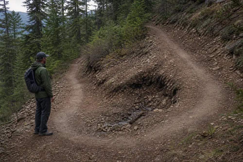 Hiker stopping before an undercut switchback with loose soil and blocked drainage - damaged switchback | KnowOutdoor