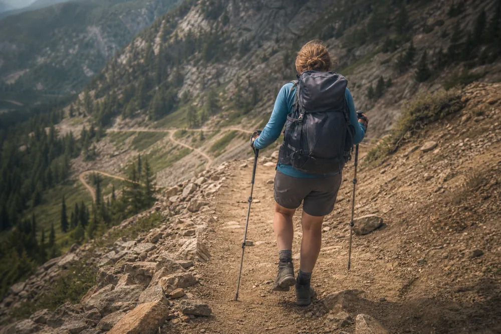 Hiker descending a straight trail section between switchbacks using trekking poles planted ahead with short controlled steps - descending switchbacks | KnowOutdoor