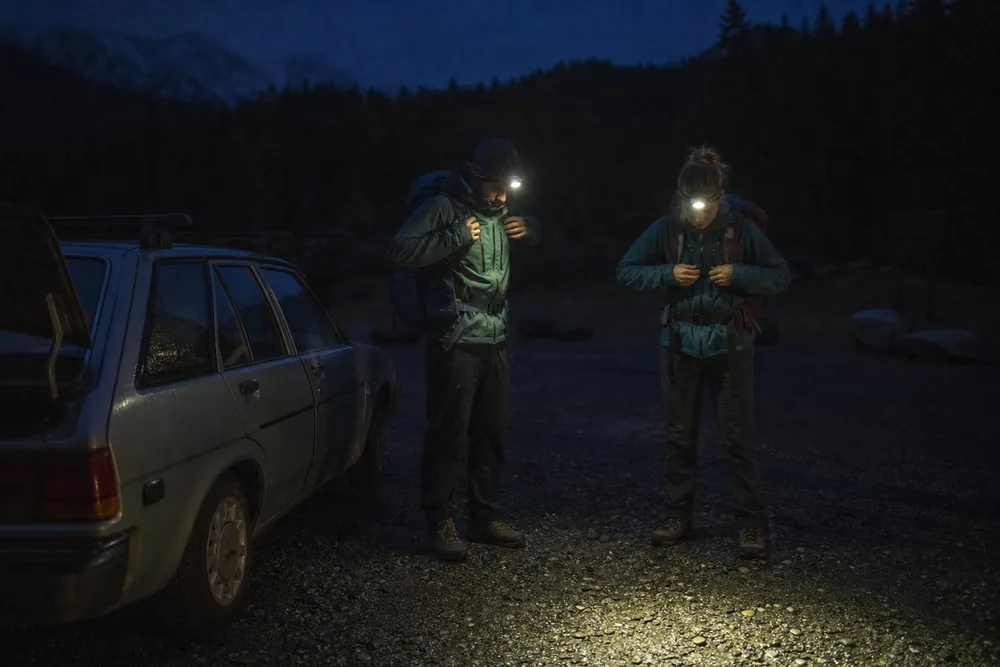 Two hikers adjusting packs beside a car at a dim gravel trailhead before dawn with headlamps switched on - sunrise and sunset hiking | KnowOutdoor