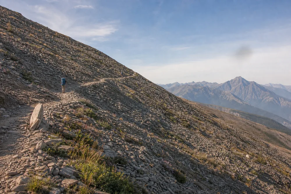 Two widely spaced hikers climbing a steep scree and tundra slope above treeline in low morning light - hiking at altitude on a day hike | KnowOutdoor