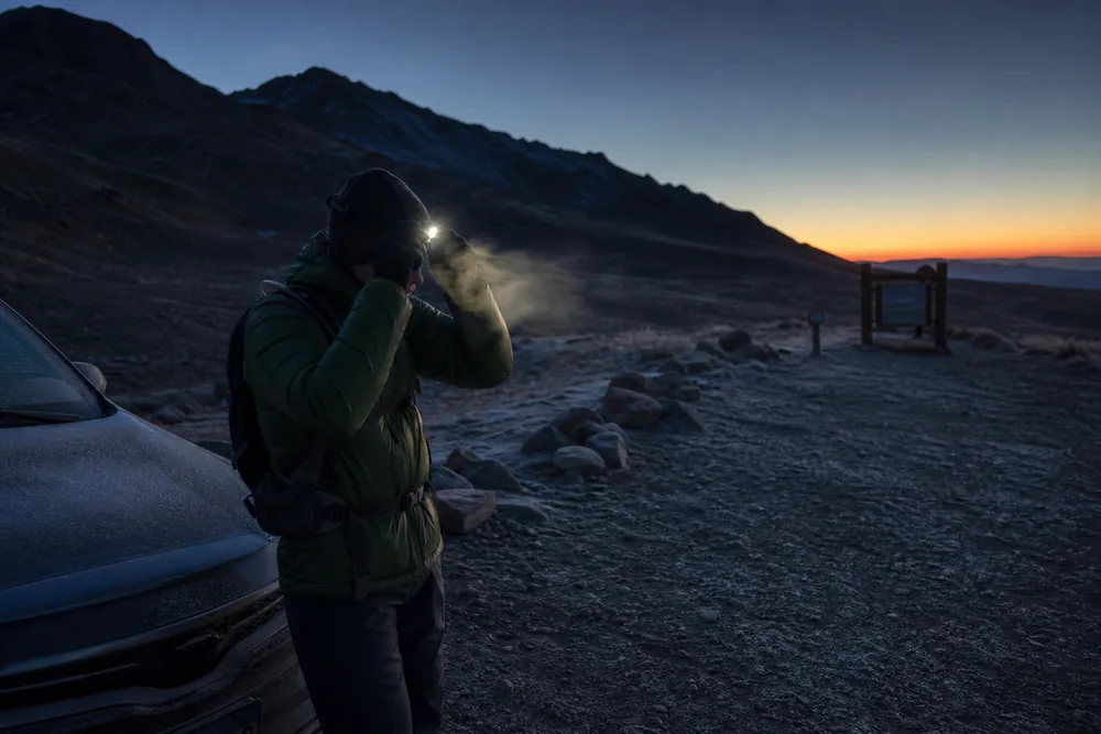 Frost-covered high alpine trailhead before dawn with a hiker adjusting a headlamp beside a car - hiking at altitude on a day hike | KnowOutdoor