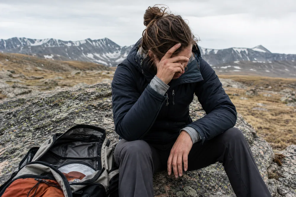 Hiker resting on a boulder on a high ridge with head lowered and one hand at the temple - hiking at altitude on a day hike | KnowOutdoor