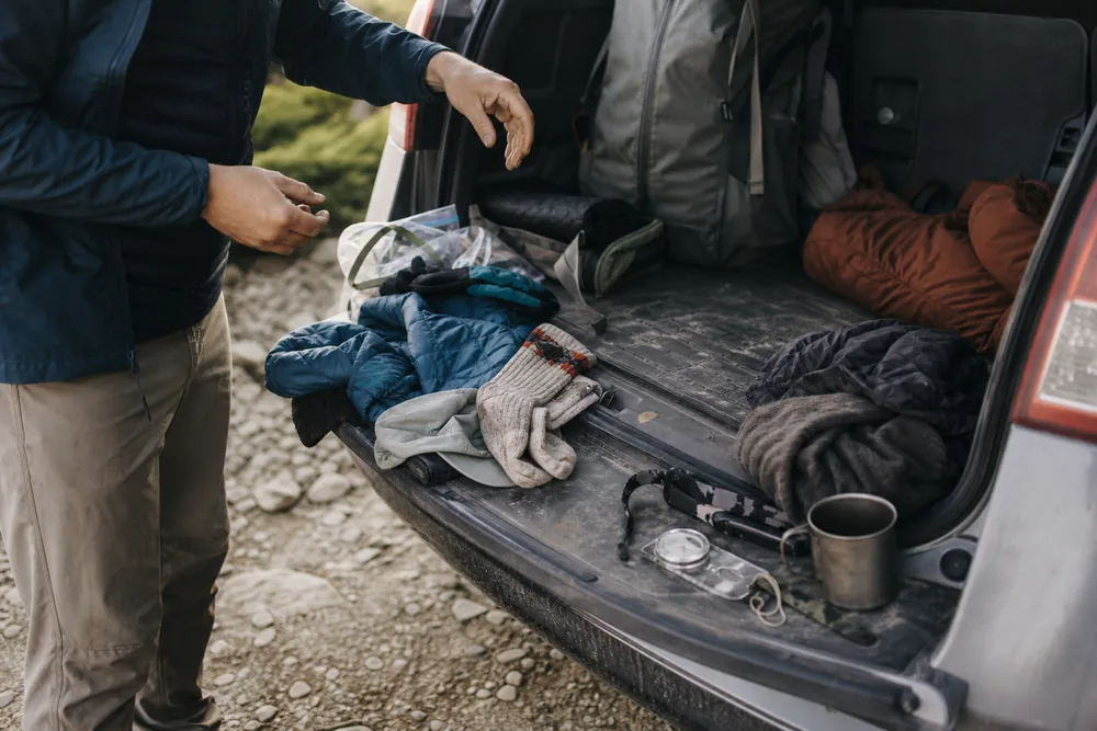 Hiking gear sorted into two piles on the open tailgate of a car at a gravel trailhead - deciding what to leave behind | KnowOutdoor