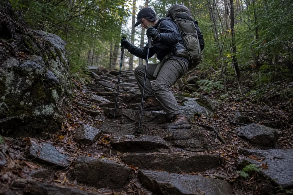 A hiker with a loaded daypack descending a steep rock staircase with both trekking poles planted on the step below - heavy pack descent technique | KnowOutdoor