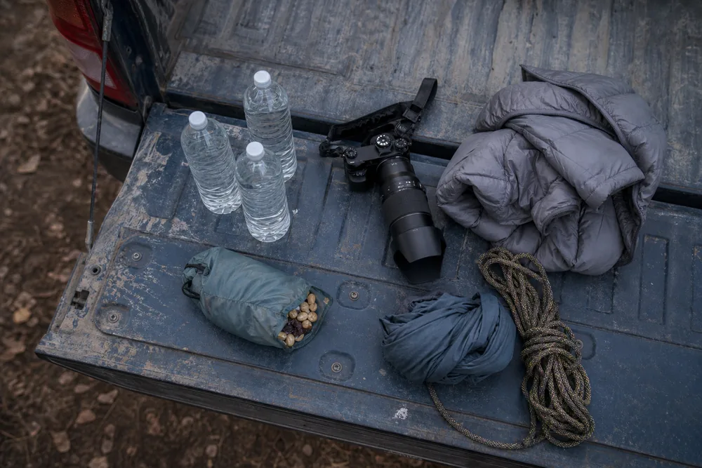 Water bottles, a camera with telephoto lens, spare layers and snacks laid out on a car tailgate before a day hike - heavy daypack loads | KnowOutdoor