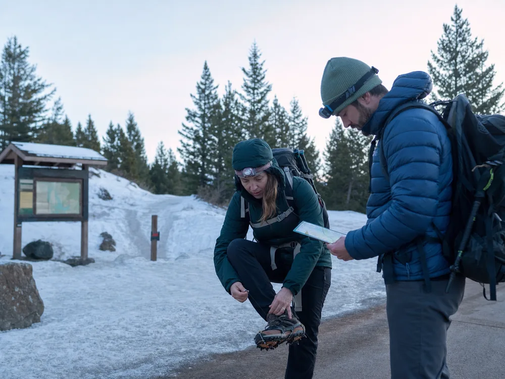 Two winter hikers checking traction headlamps insulated layers and an offline route beside a snowy trailhead board - winter hiking trailhead check | KnowOutdoor