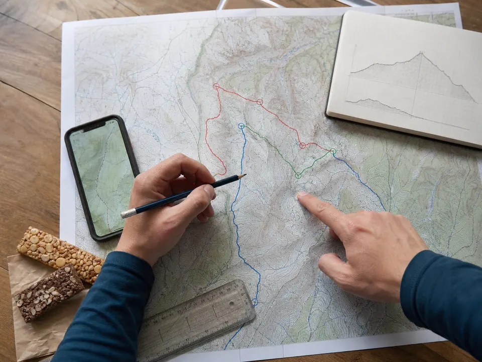 Hands comparing a paper trekking map, route marks, elevation notes, and offline map before a first trek