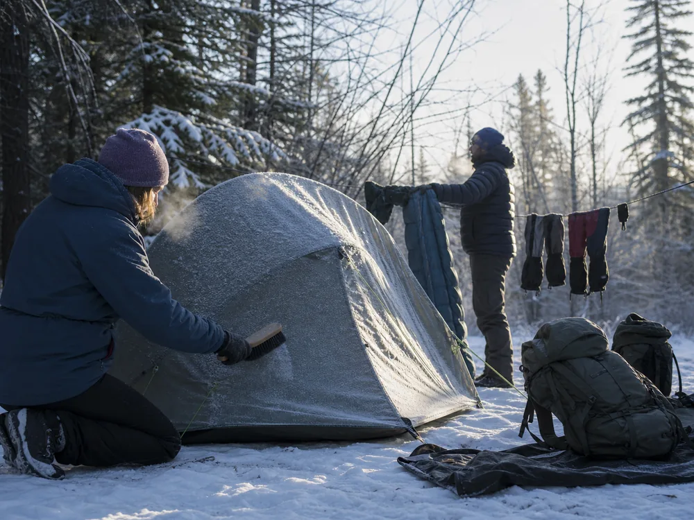 Winter trekkers brushing frost from a shelter and airing damp gear outside the dry sleeping area - multi-day frost management | KnowOutdoor