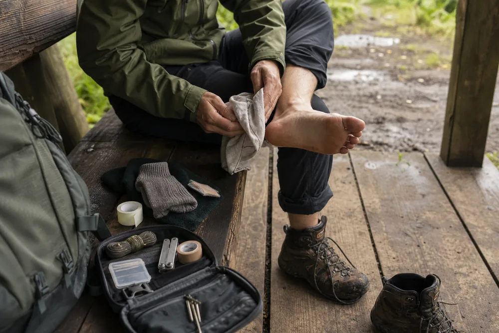Trekker drying a foot and organizing blister tape, spare socks, cord, and repair tools beside a pack